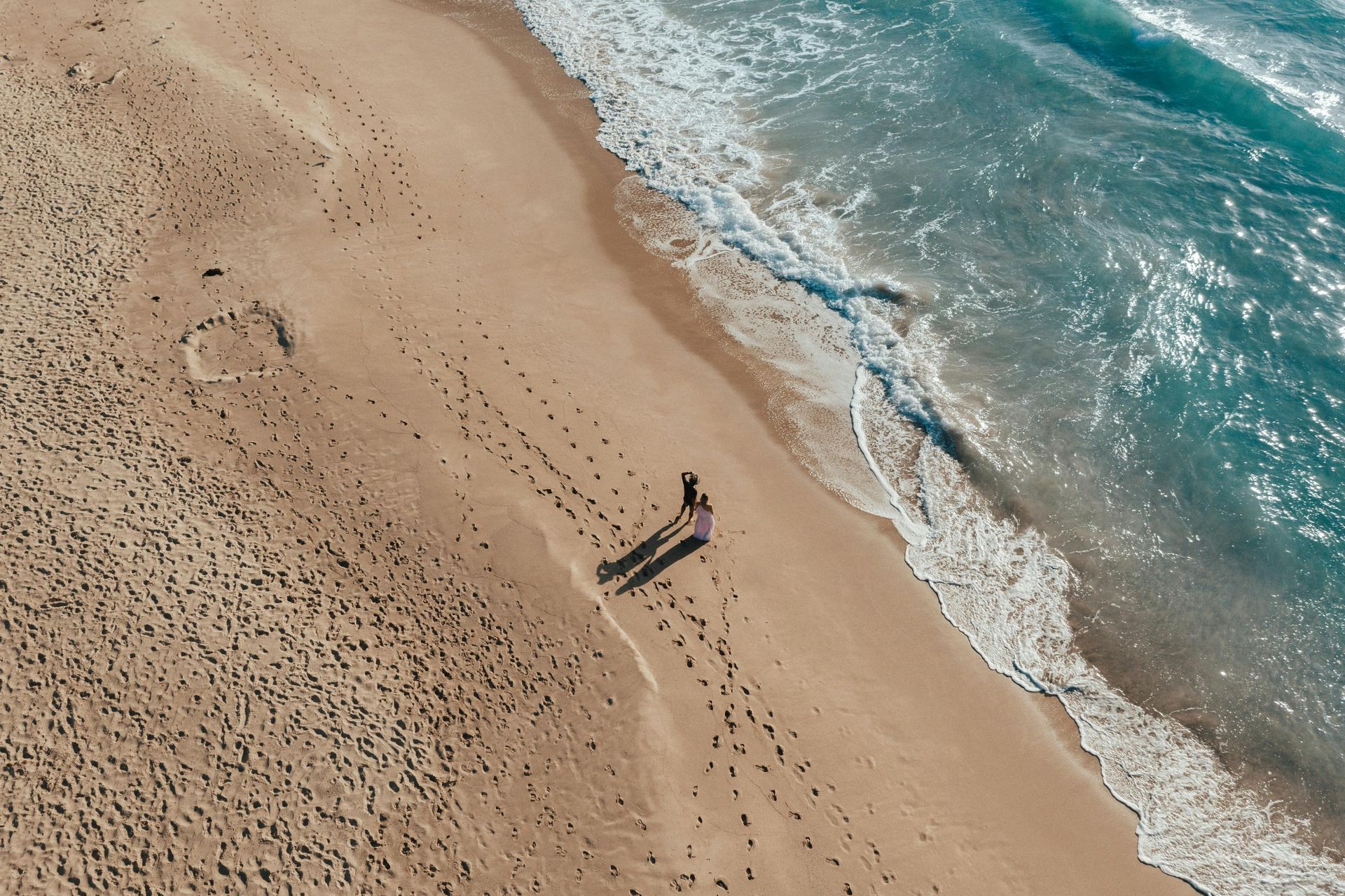 An aerial view of two people walking along a sandy beach beside turquoise ocean waves.