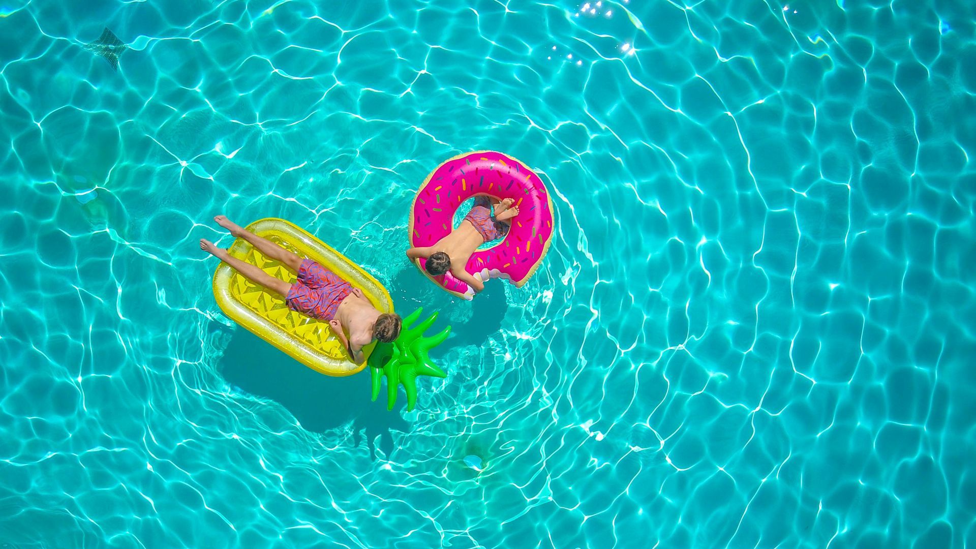 Two people float in a blue swimming pool on a yellow pineapple-shaped float and a bright pink circular inner tube.