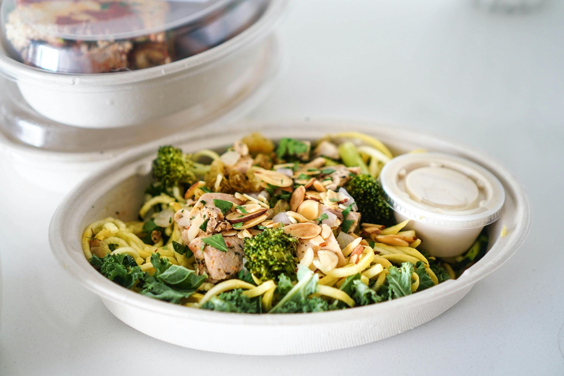 A salad bowl with chicken, broccoli, kale, and slivered almonds, accompanied by a small side of dressing.