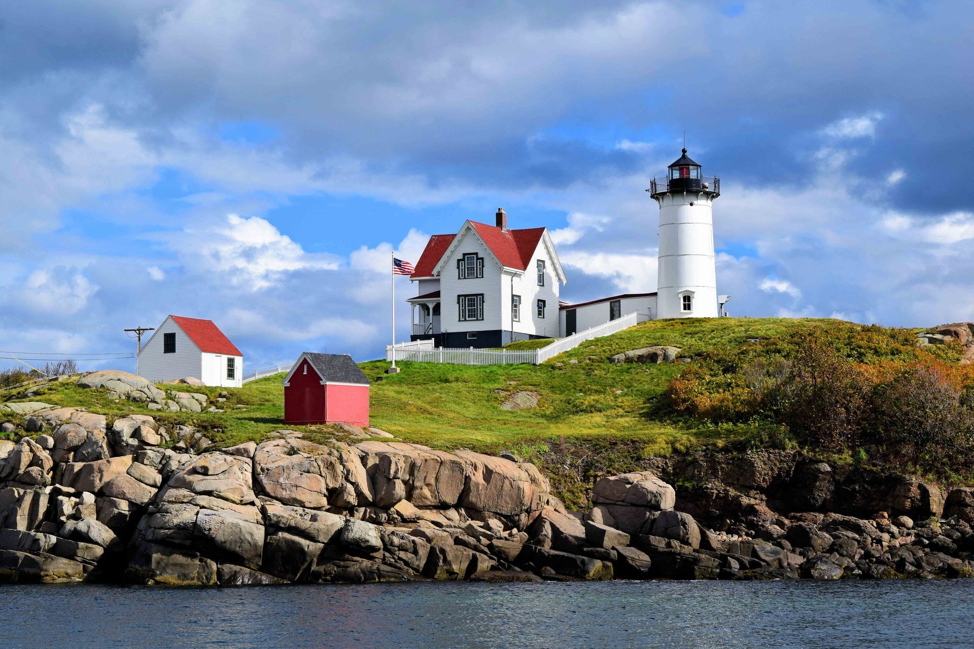 Cape Neddick Lighthouse with white buildings and a red-roofed house on a rocky, grassy island under a cloudy blue sky.