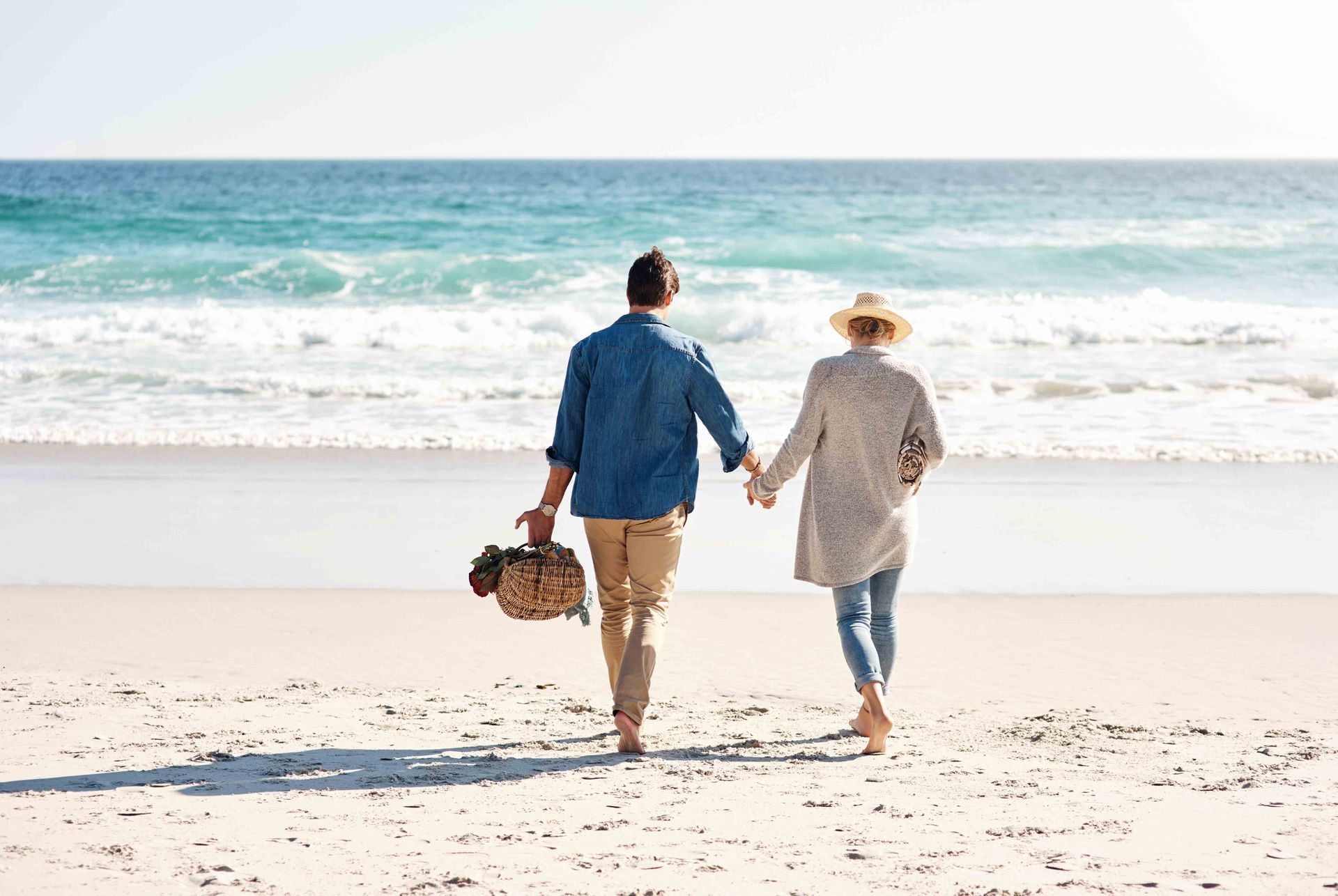 A couple walks hand-in-hand along a sunny beach, carrying a picnic basket toward the ocean.