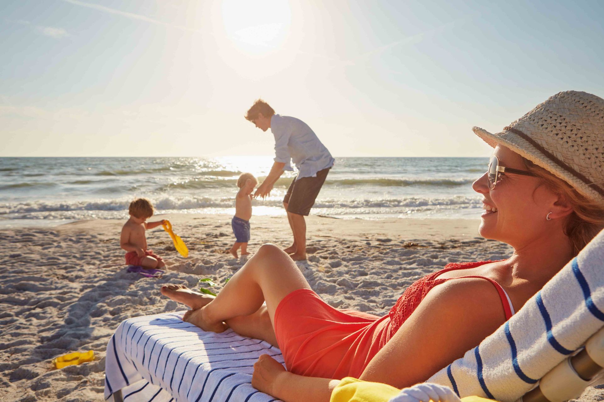 A smiling person wearing a straw hat rests on a beach chair, watching a family play in the surf under bright sunlight.