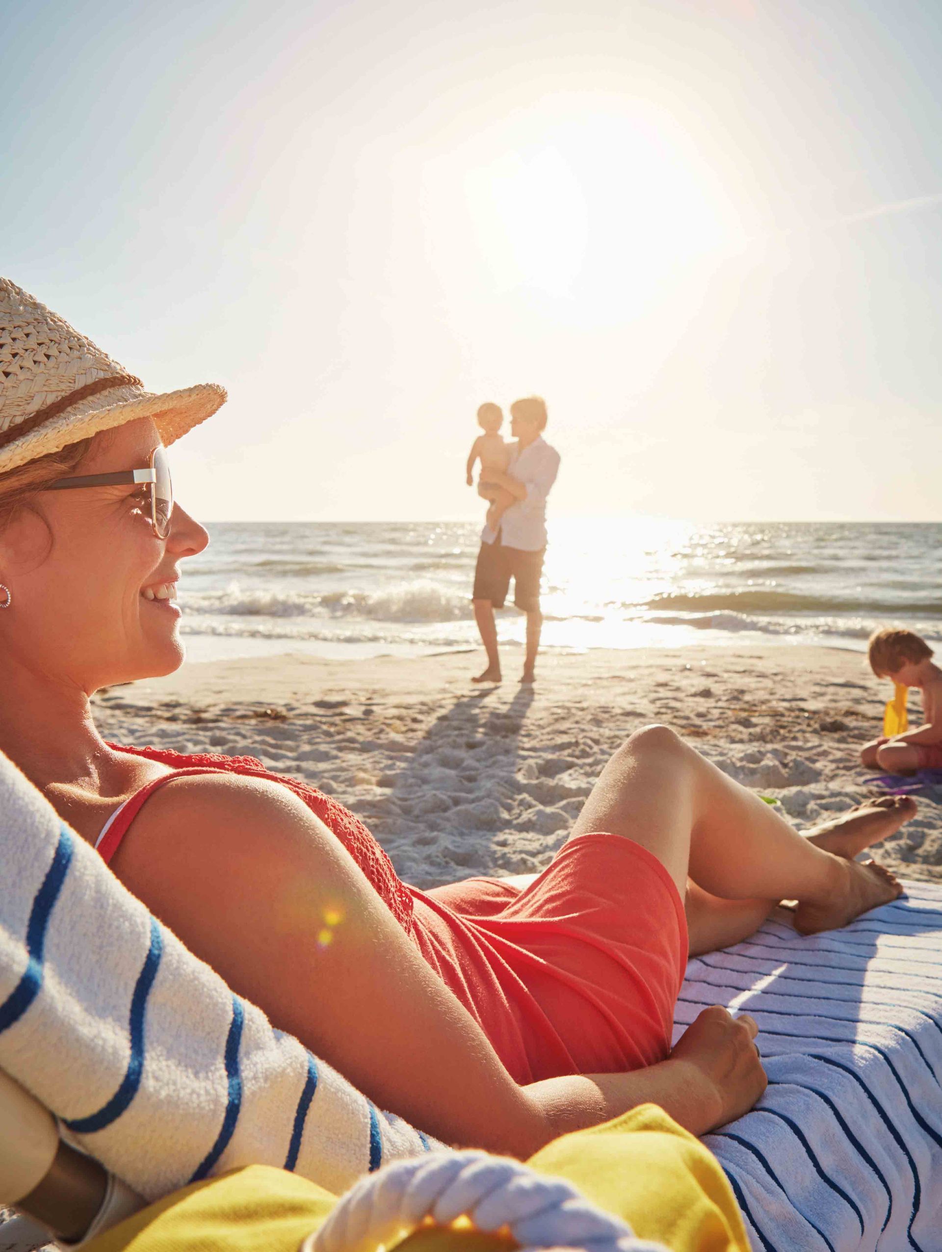 A person wearing a sun hat lounges on a beach towel while others play on the sunny shoreline in the background.