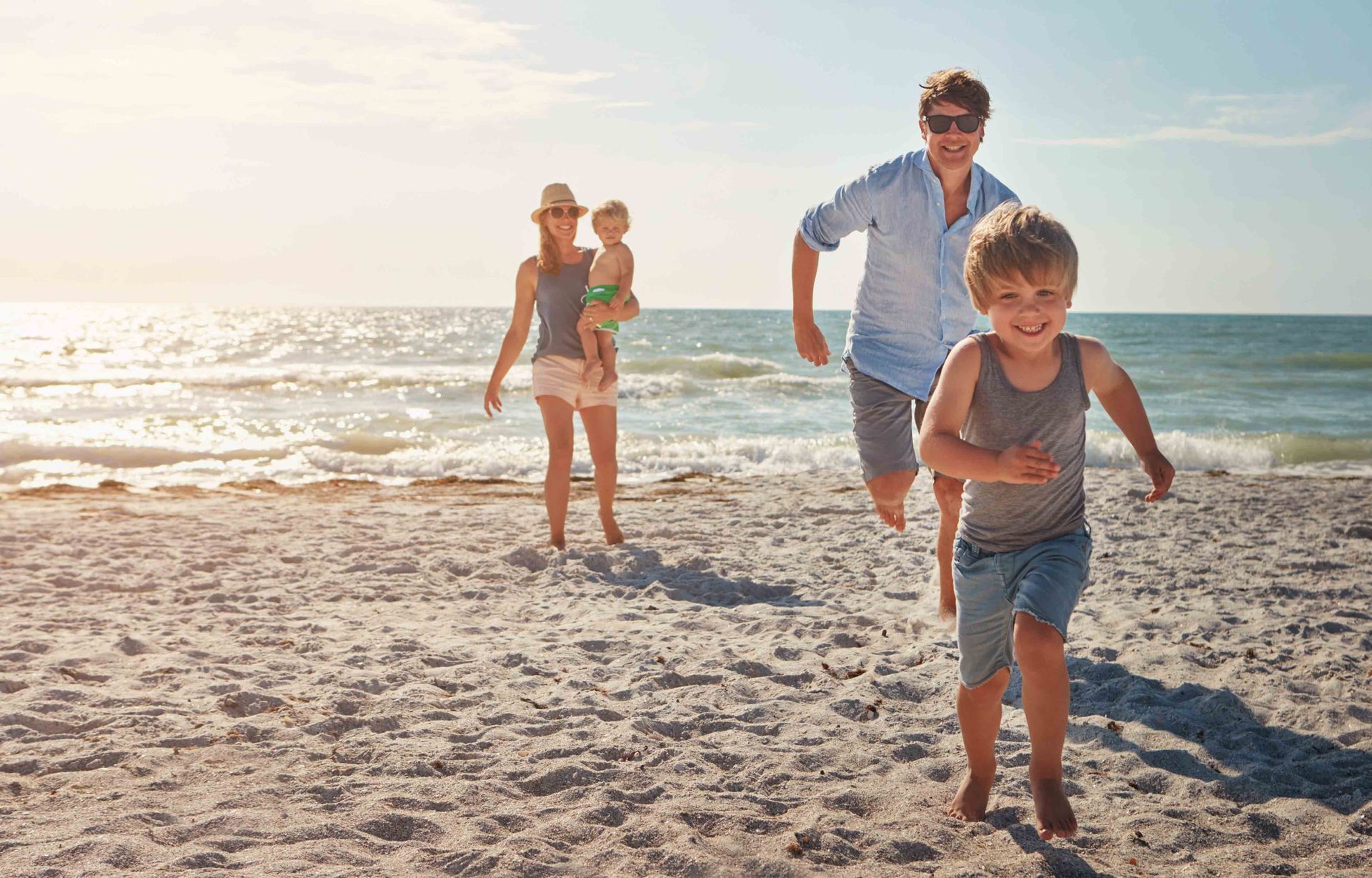 A family runs and plays together on a sunlit beach with the ocean in the background.