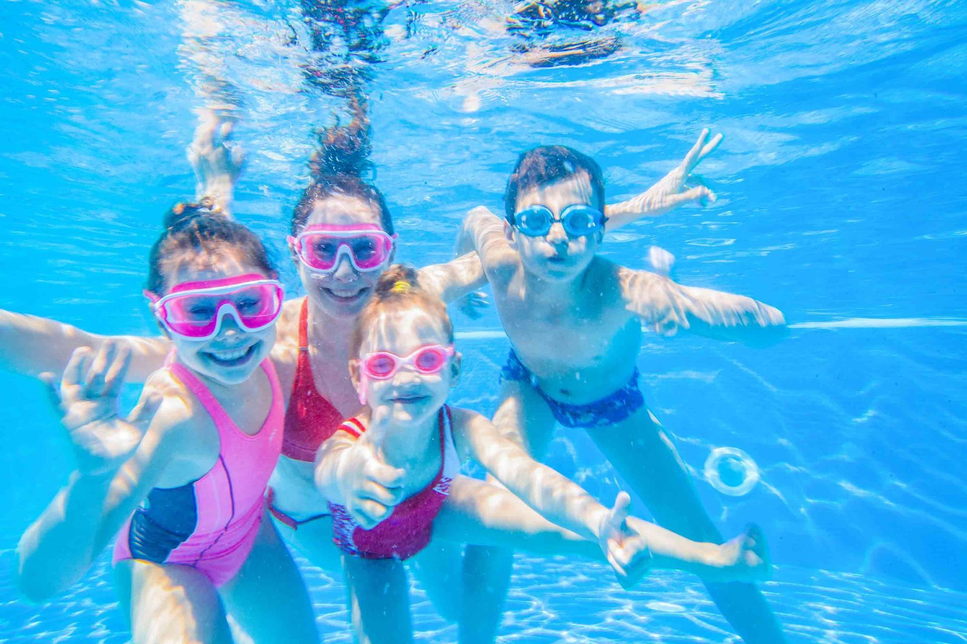 Four people wearing swimsuits and goggles pose underwater in a pool, smiling and waving at the camera.
