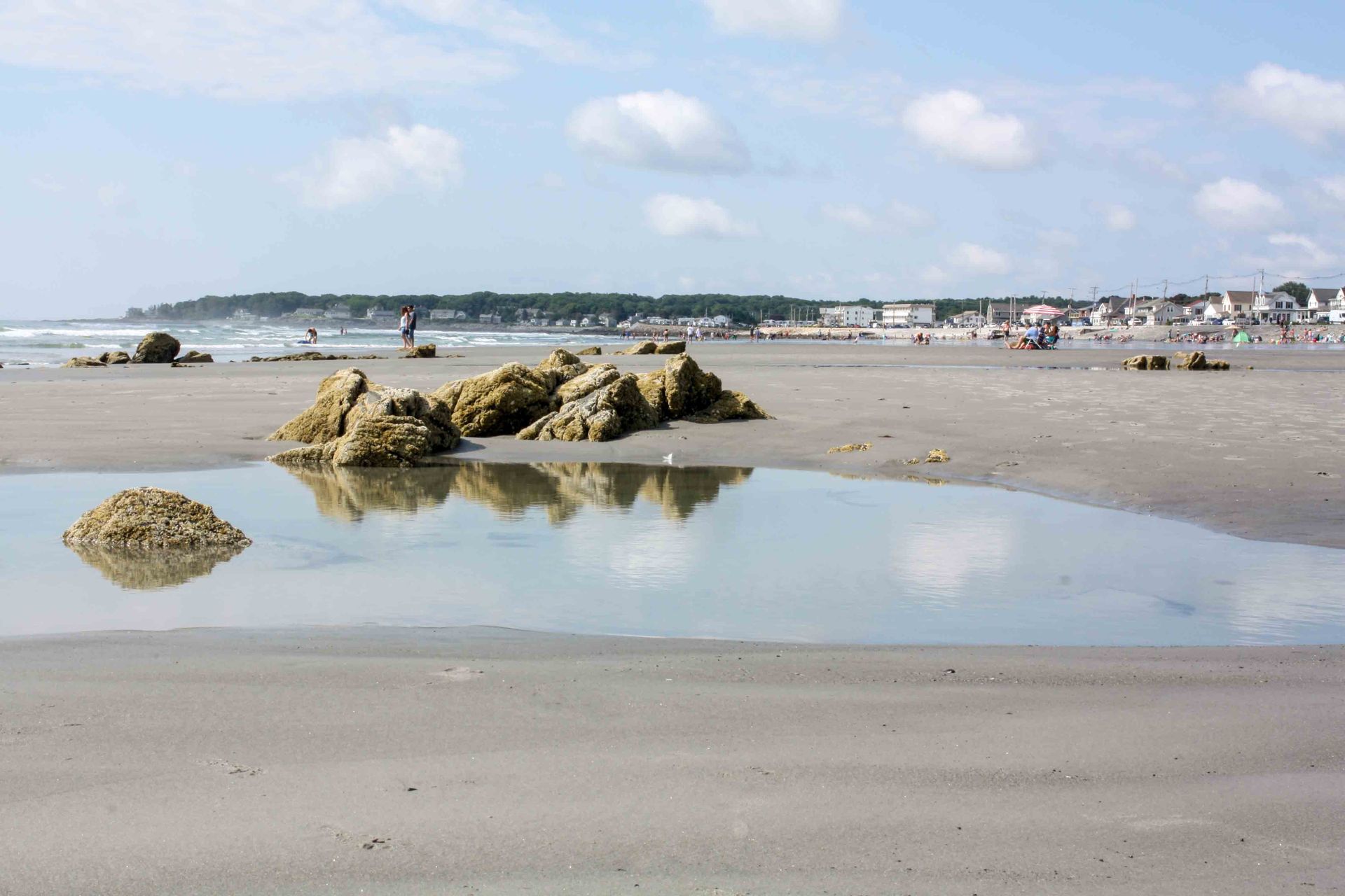 Large rocks on a sandy beach sit by a tide pool reflecting the sky, with distant buildings along the shoreline.