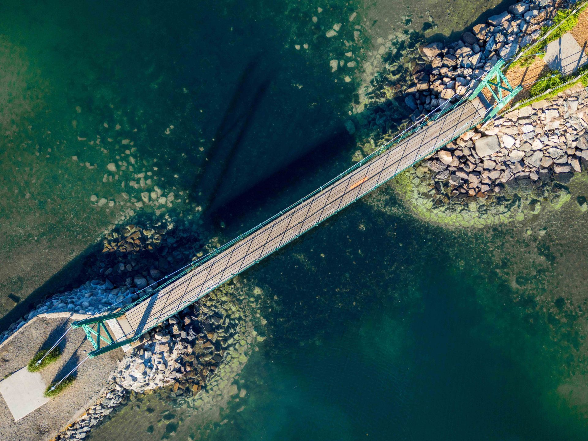 A high-angle aerial view of a narrow pedestrian suspension bridge crossing over a clear, dark blue body of water.