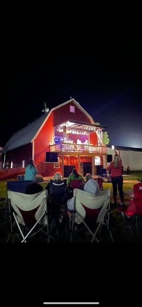 A group of people are sitting in chairs in front of a red barn at night.