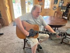 A man is sitting in a chair playing an acoustic guitar in a living room.