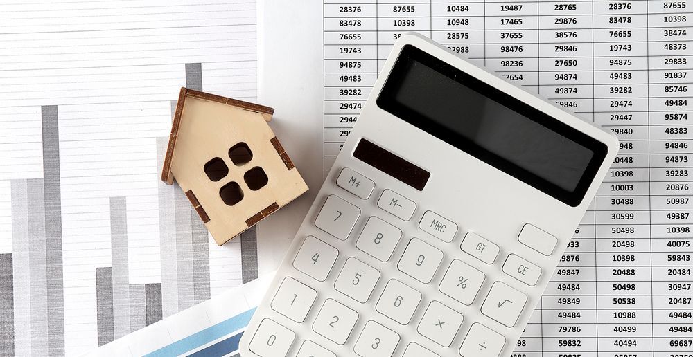 Calculator and wooden house icon on financial charts, symbolizing budgeting or home finance.