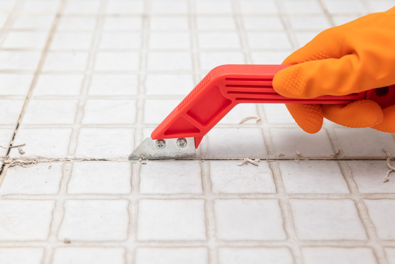 Hand in orange glove using a red grout saw to remove grout from between white tiles.