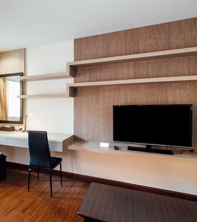 A modern hotel room with a desk, TV, and shelves on a wood-paneled wall, set against a window and wood floor.