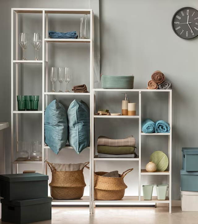White shelving unit with household items: pillows, towels, baskets, glasses, and decorative objects.
