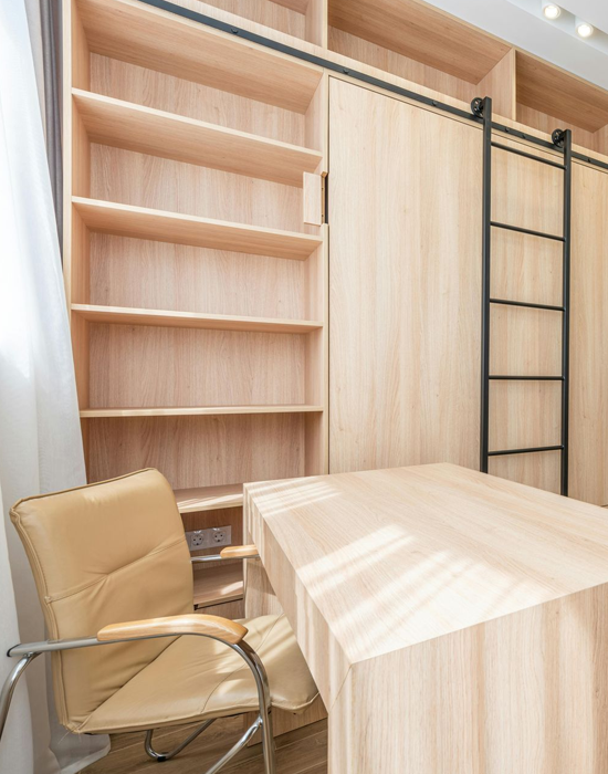 Light wood home office with desk, shelving, sliding door, ladder, and a beige office chair.