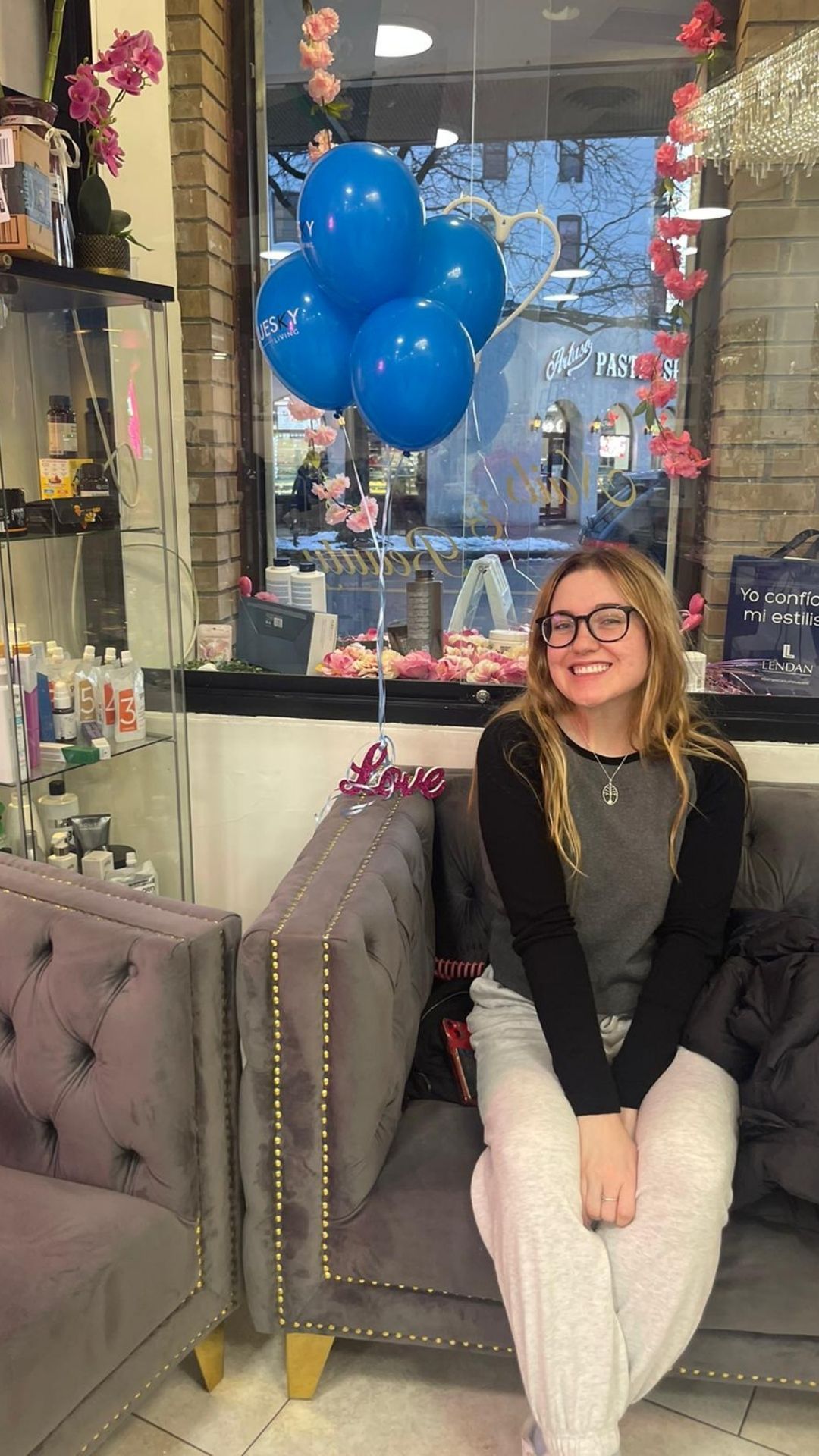 A woman is sitting on a couch in front of a window with blue balloons.