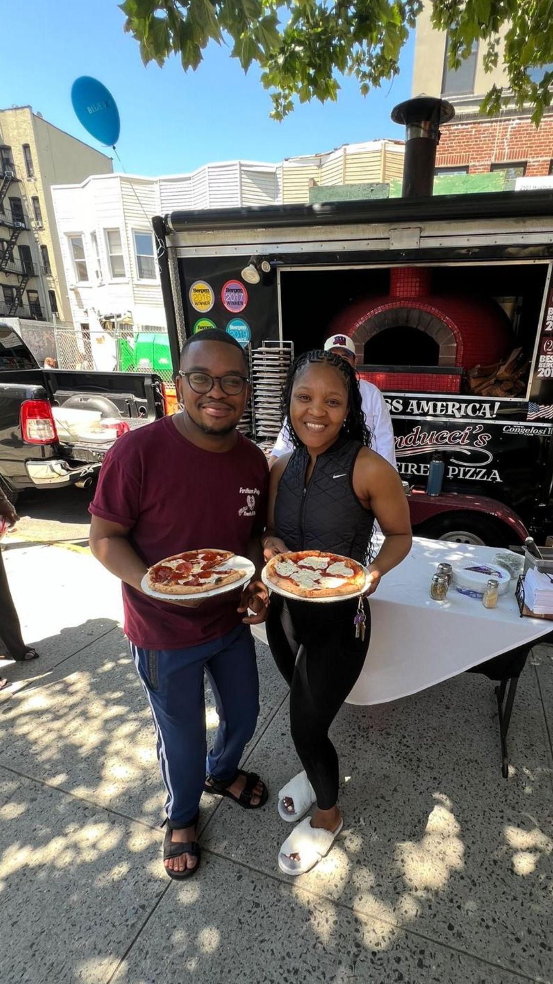 A man and a woman are holding pizzas in front of a pizza truck.