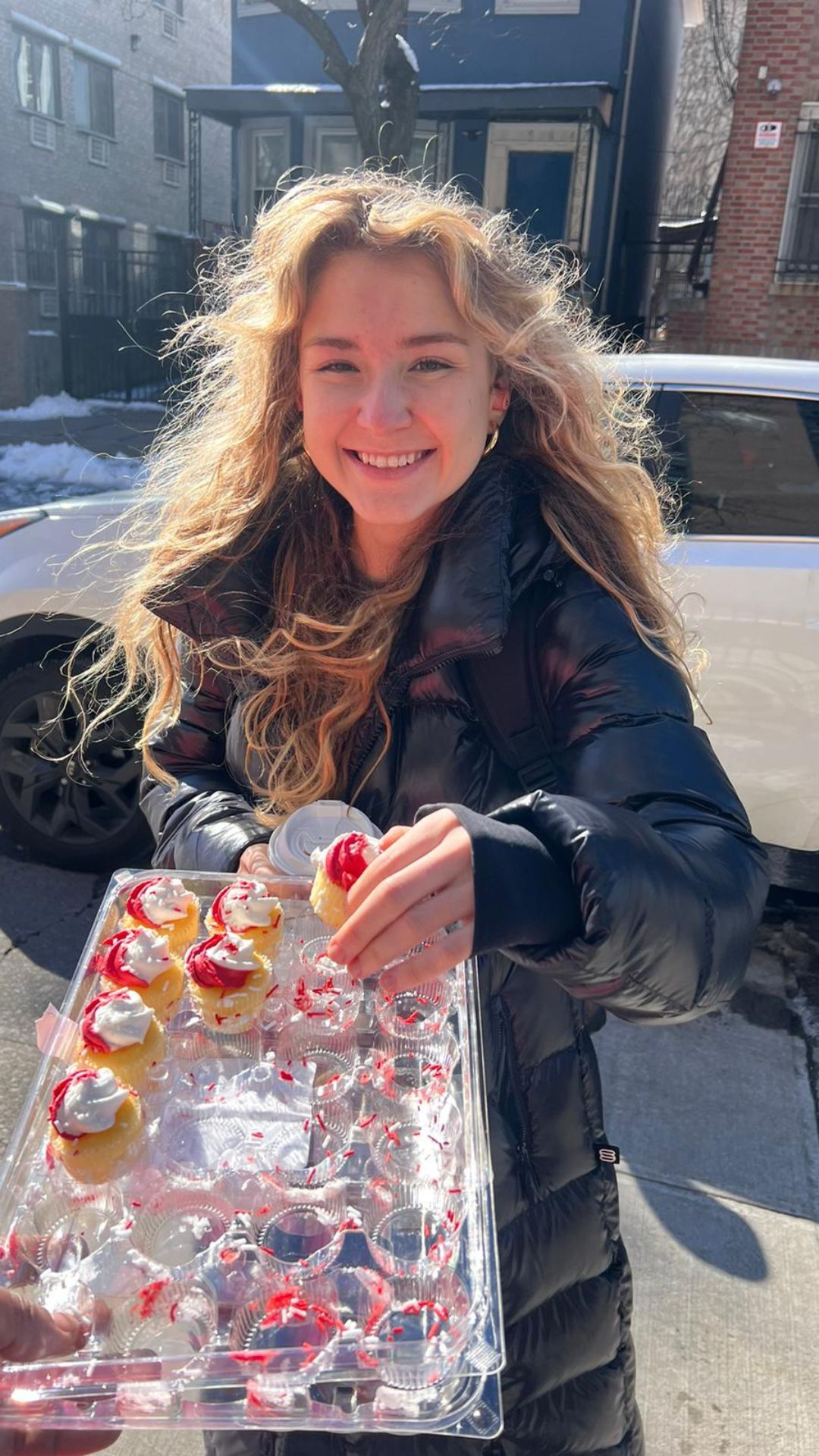 A woman is holding a tray of cupcakes and smiling.