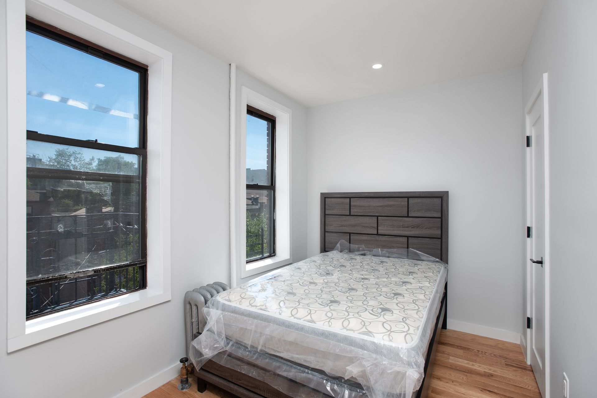 A bedroom with a bed , two windows and a mattress wrapped in plastic.