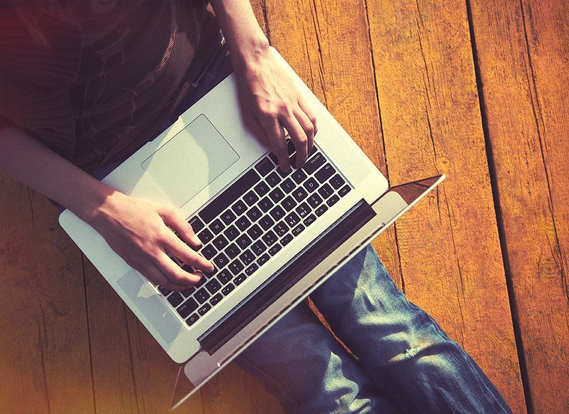 A person sits on a wooden floor, typing on an open silver laptop viewed from directly above.