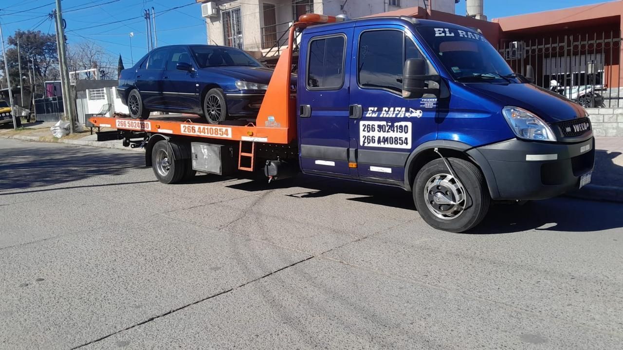 Grúa azul que transporta un coche azul en una calle de la ciudad. Plataforma de remolque naranja.