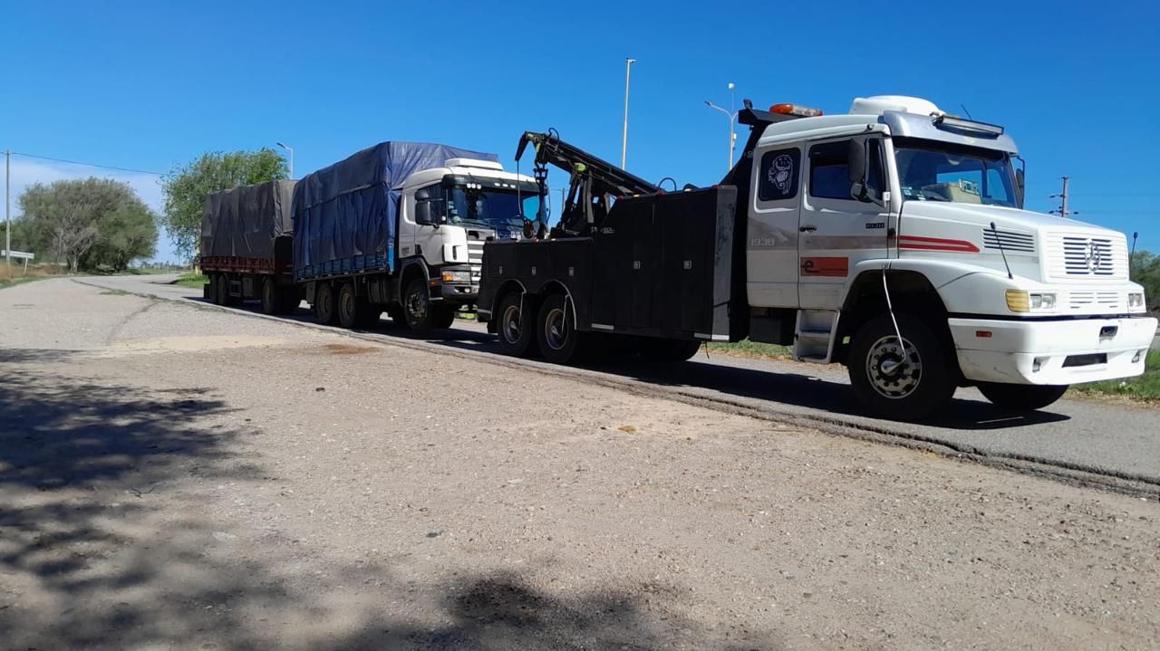 Grúa remolcando un semirremolque con un remolque cubierto con una lona azul en una carretera soleada.