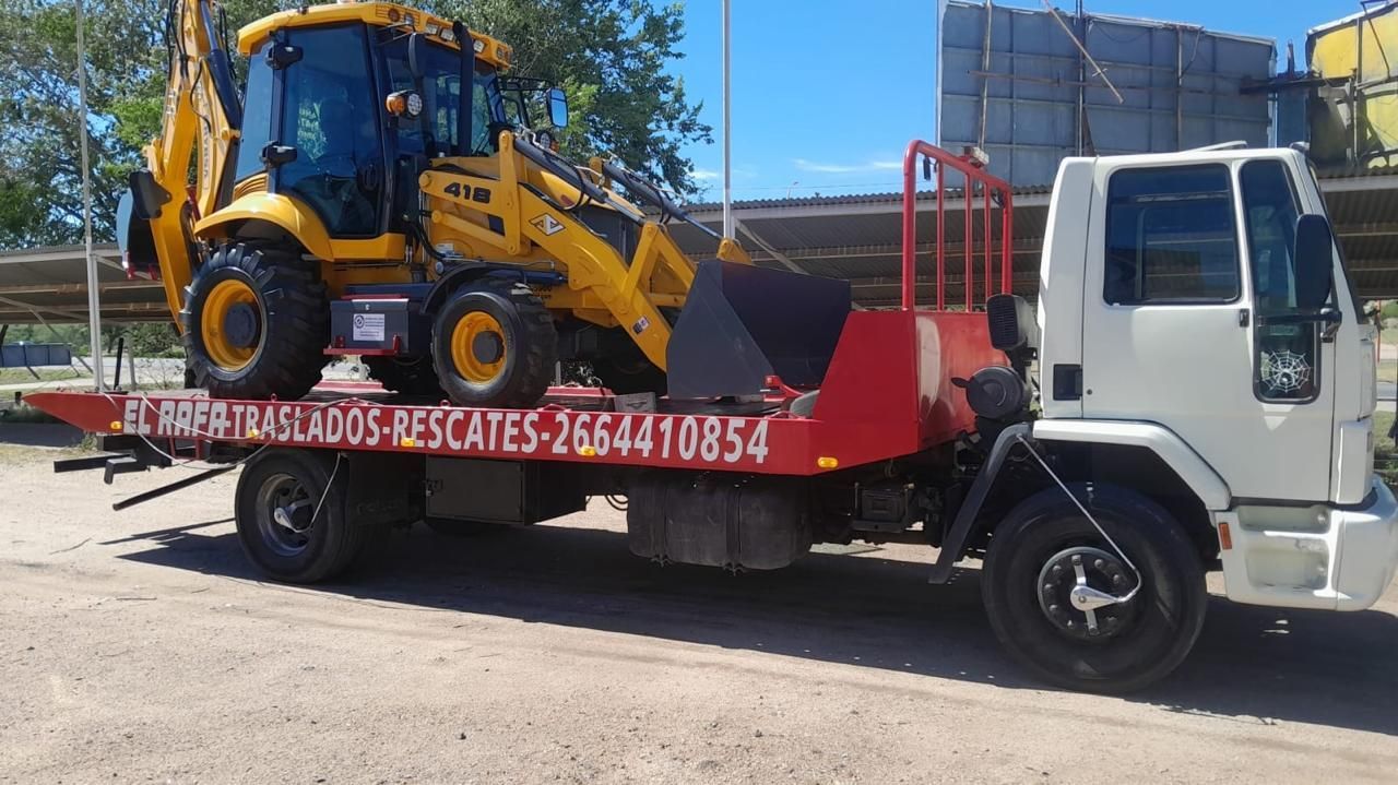 Retroexcavadora amarilla sobre una grúa de plataforma. El camión es blanco y rojo. Al aire libre, soleado.