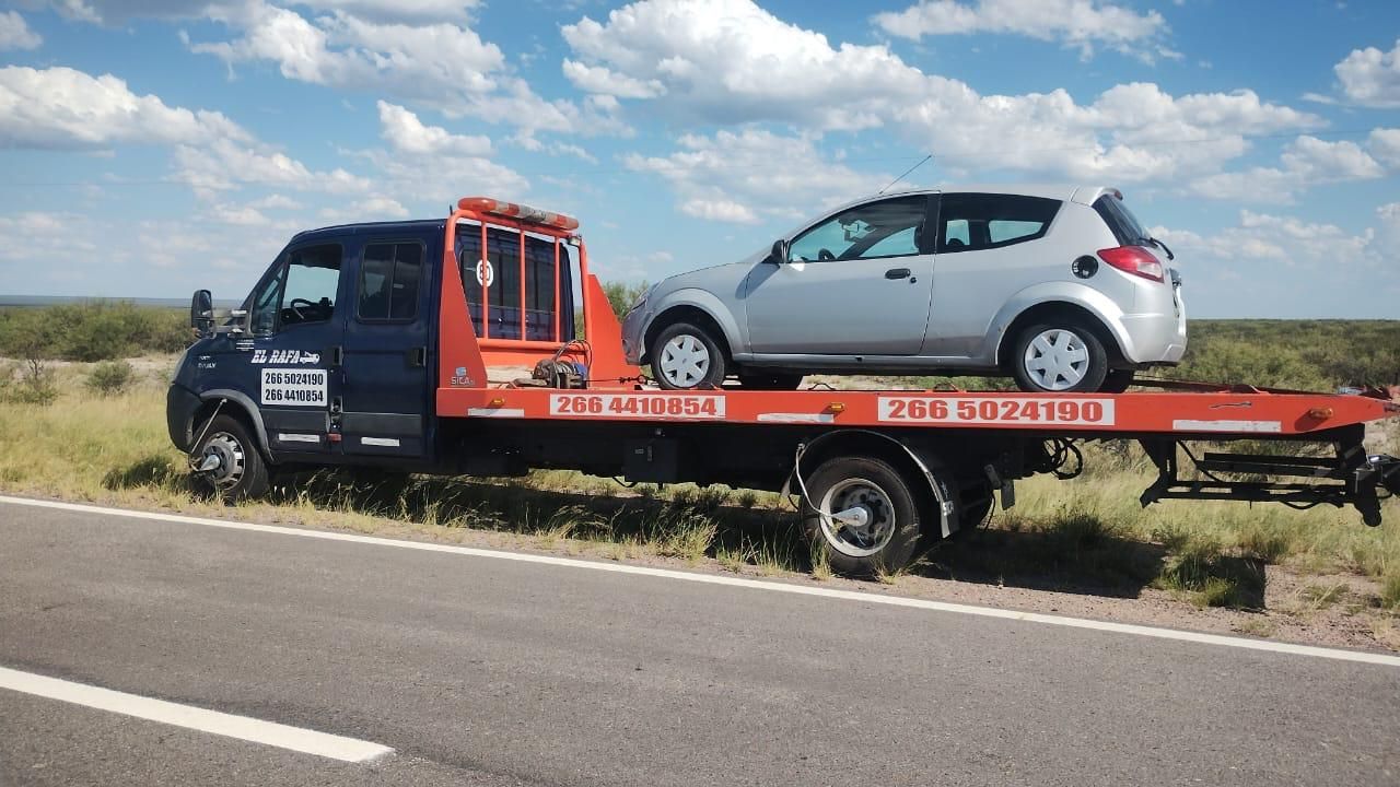 Grúa con coche plateado sobre la plataforma, aparcada al borde de la carretera, bajo un cielo azul.