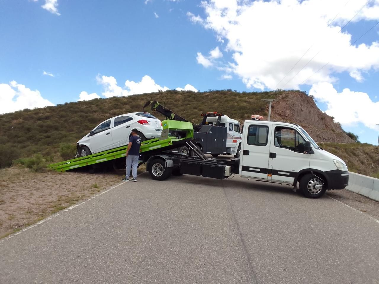 Un coche blanco está siendo cargado en una grúa verde en una carretera; una persona está de pie cerca.