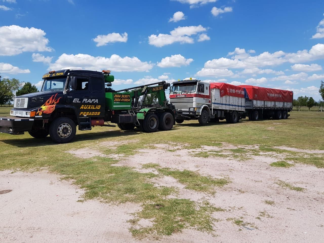 Grúa remolcando un camión con remolque cubierto en un campo de hierba, bajo un cielo azul.