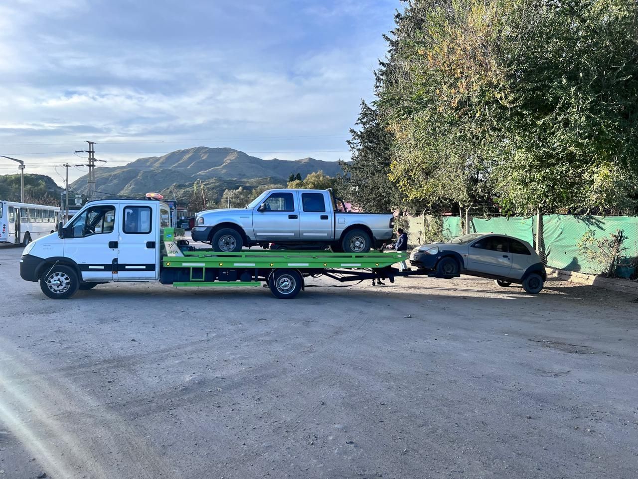Grúa que transporta una camioneta pickup y remolca un automóvil más pequeño en un terreno de grava cerca de las montañas.