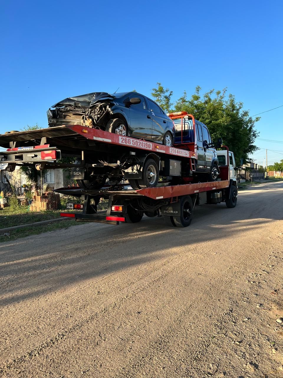 Grúa que transporta un coche negro muy dañado por un camino de tierra. Cielo despejado, día soleado.