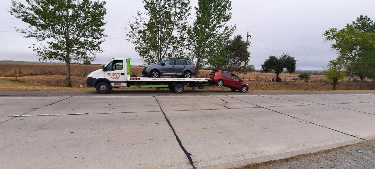 Una grúa transporta un todoterreno gris y un coche rojo por una carretera gris. Al fondo se ve un campo con árboles.