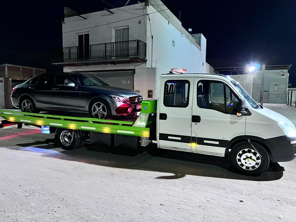 Grúa de noche cargando un coche plateado. Edificio al fondo, plataforma de carga verde.