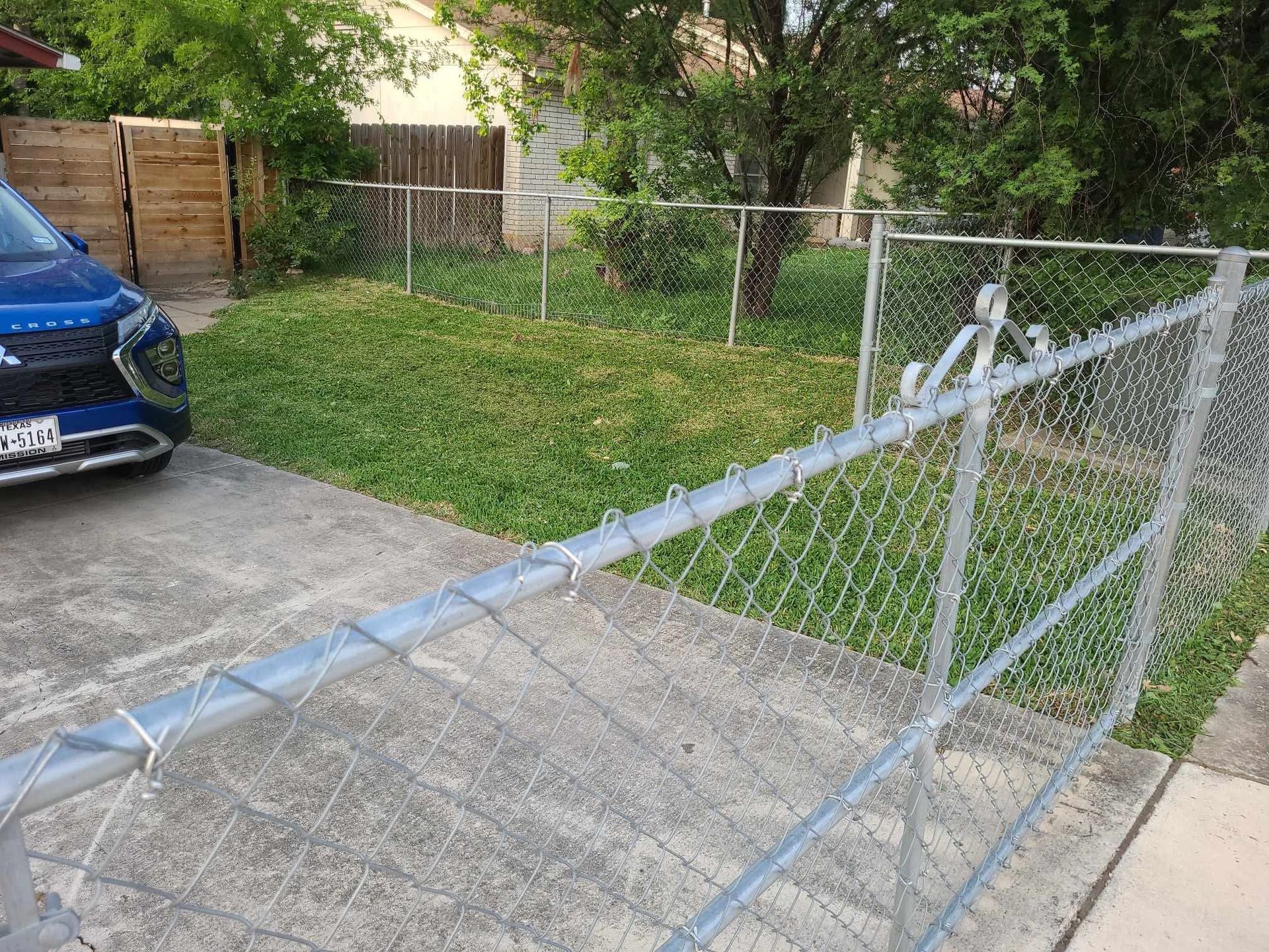 Chain-link fence and open gate across a driveway beside a blue car and grassy yard.