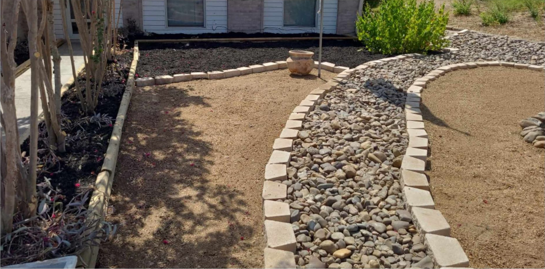 Narrow side yard beside a beige building, with a leafy tree, fence, mulch, and stacked boards.