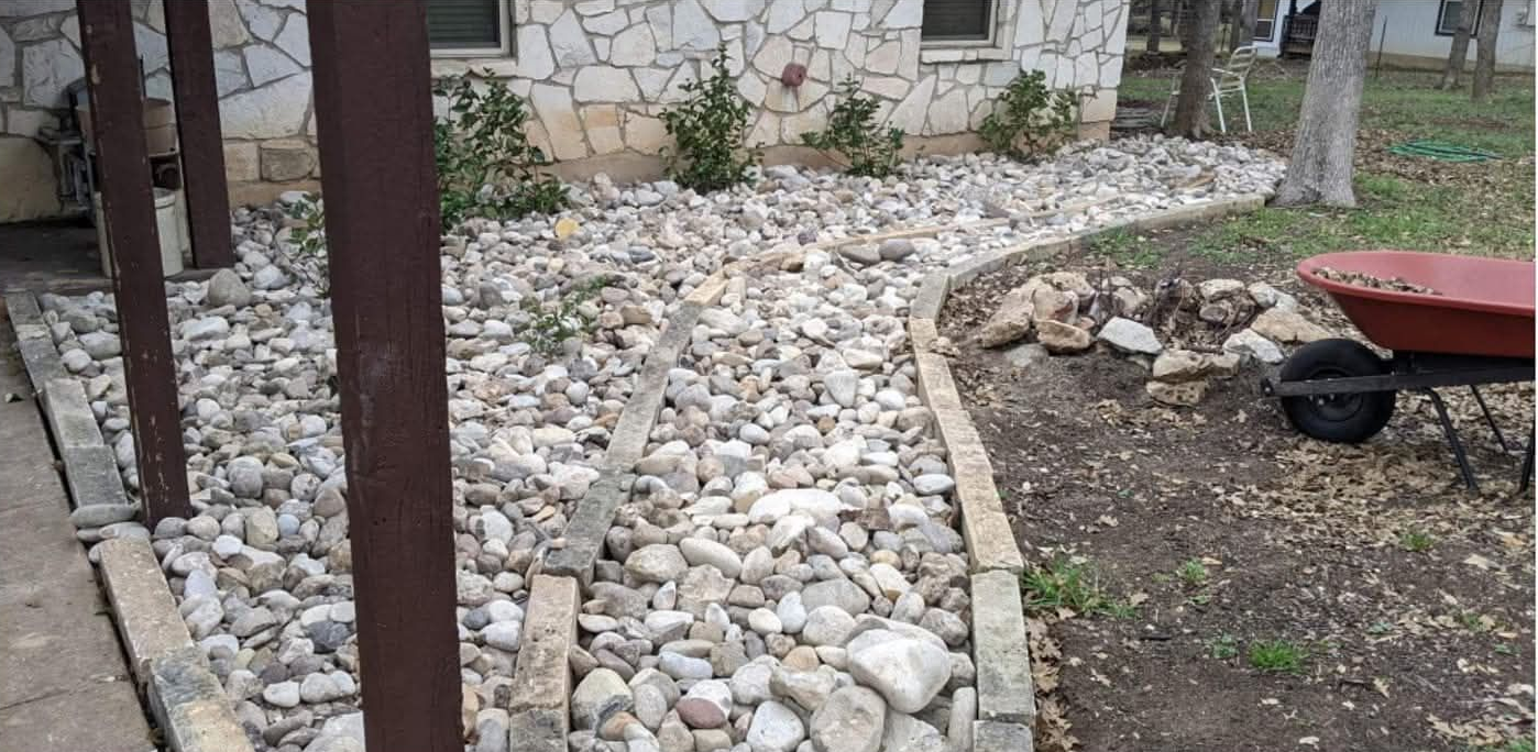Narrow side yard beside a beige building, with a leafy tree, fence, mulch, and stacked boards.