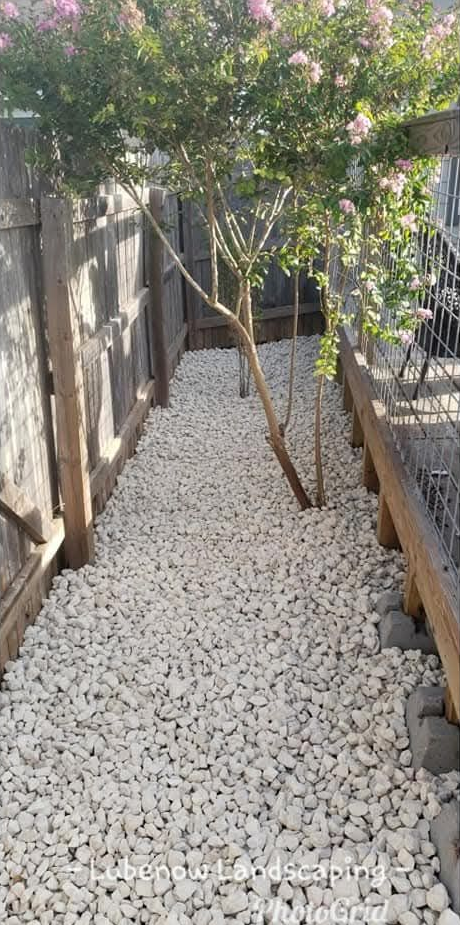 Narrow side yard beside a beige building, with a leafy tree, fence, mulch, and stacked boards.