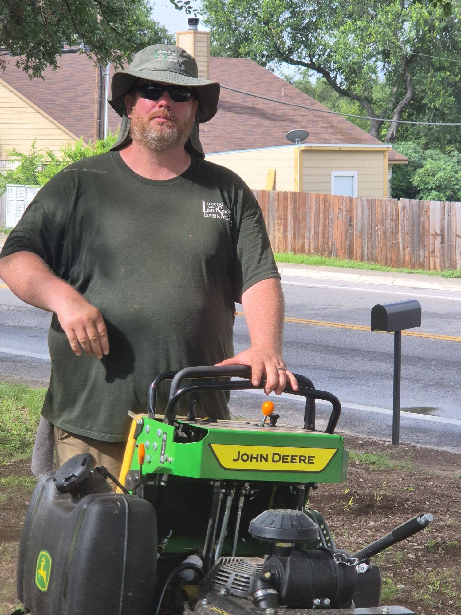 Man in black hat stands beside a green John Deere mower on a suburban street