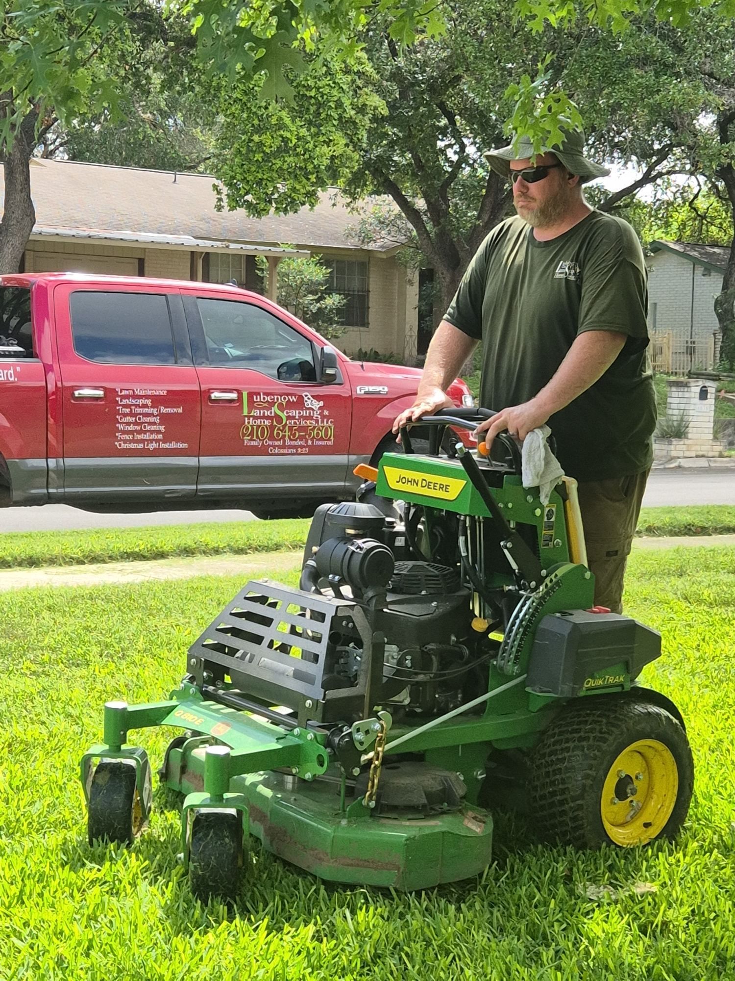 Man mowing a lawn with a green riding mower under a tree, with a red truck parked behind him.