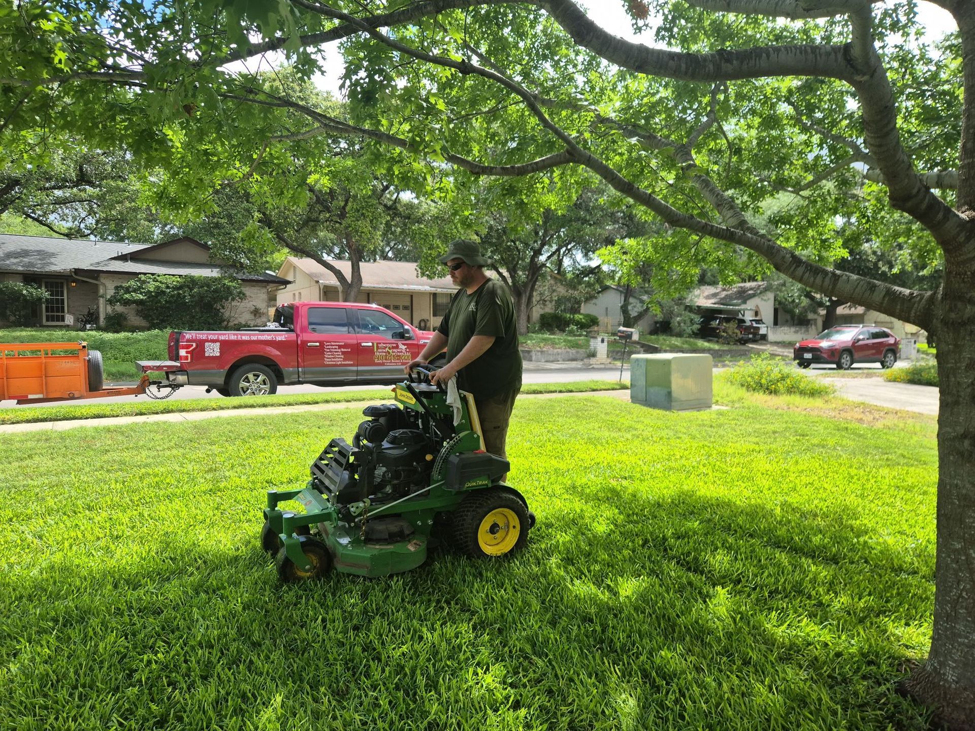 Person mowing a bright green lawn with a riding mower under trees in a suburban yard