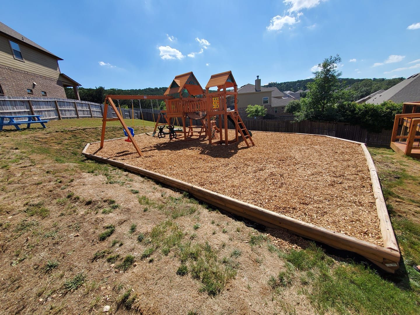 Playground with wooden climbing structure, swing set, and wood chips in a sunny fenced yard