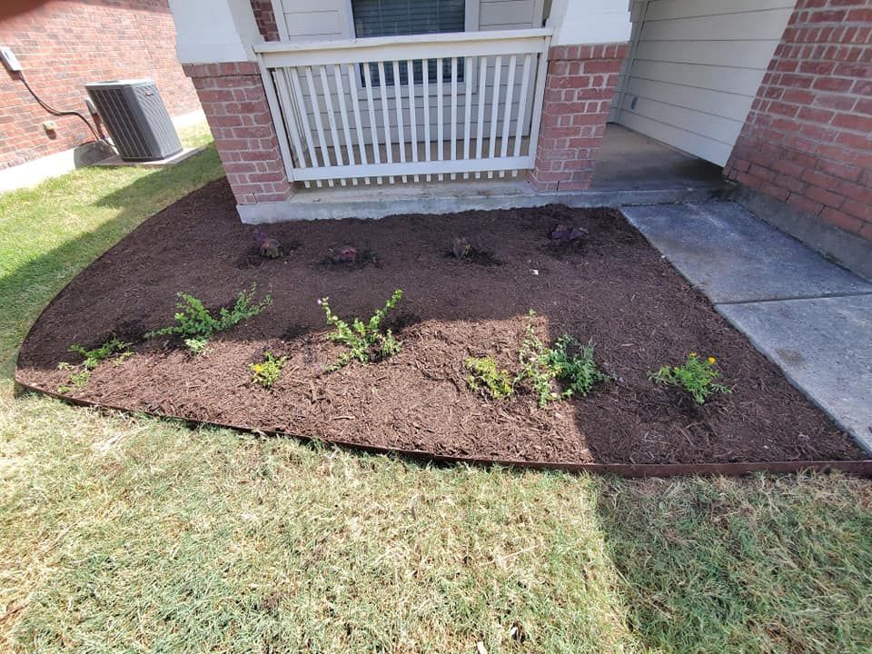 Freshly planted garden bed with small green plants beside a brick house and porch railing