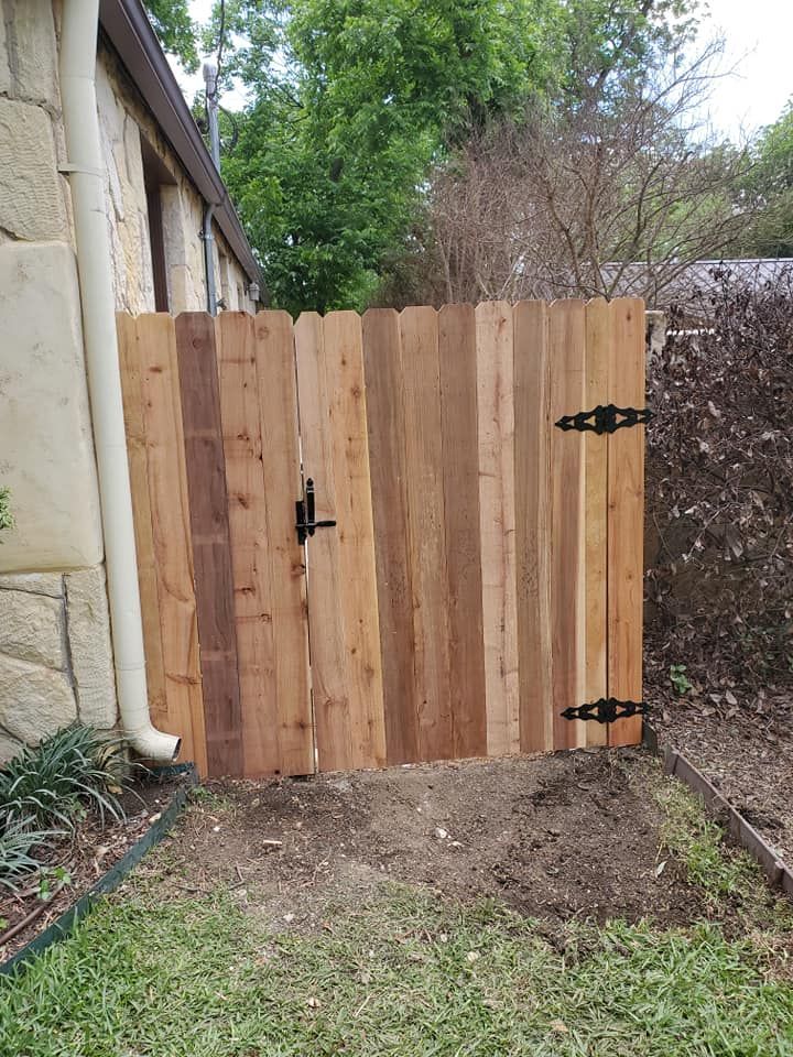 Wooden backyard gate with black hinges beside a stone house and fence, opening onto grass and dirt.