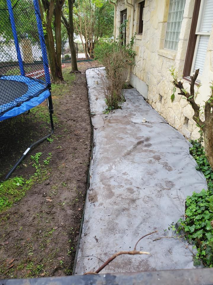 Narrow concrete path beside a house, with a trampoline and trees along the left side.