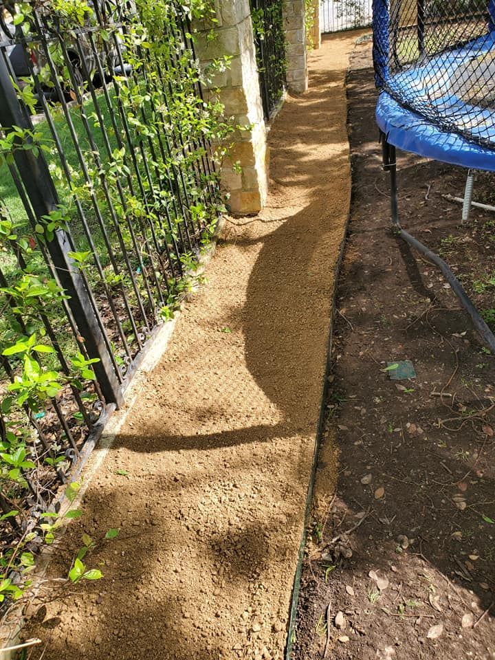 Narrow dirt path beside a metal fence and green plants, with a trampoline on the right.