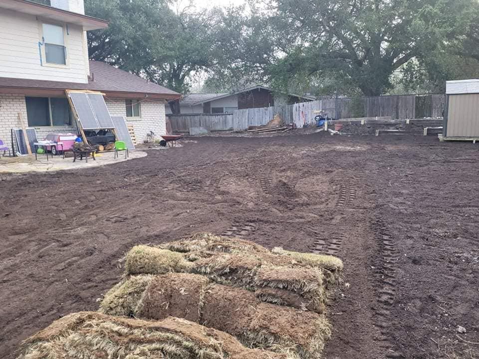 Freshly leveled dirt yard beside a house, with cut sod piled in foreground and trees and sheds in back.