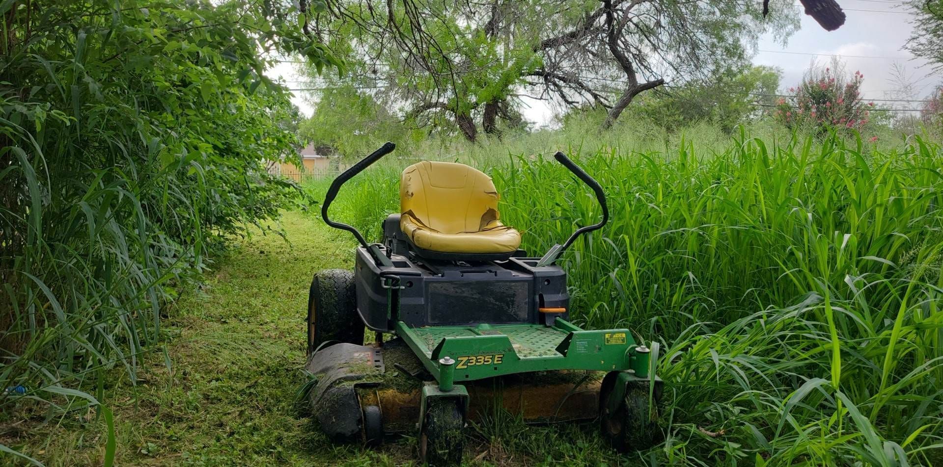 Green lawn mower with yellow seat cutting tall grass on a narrow overgrown path