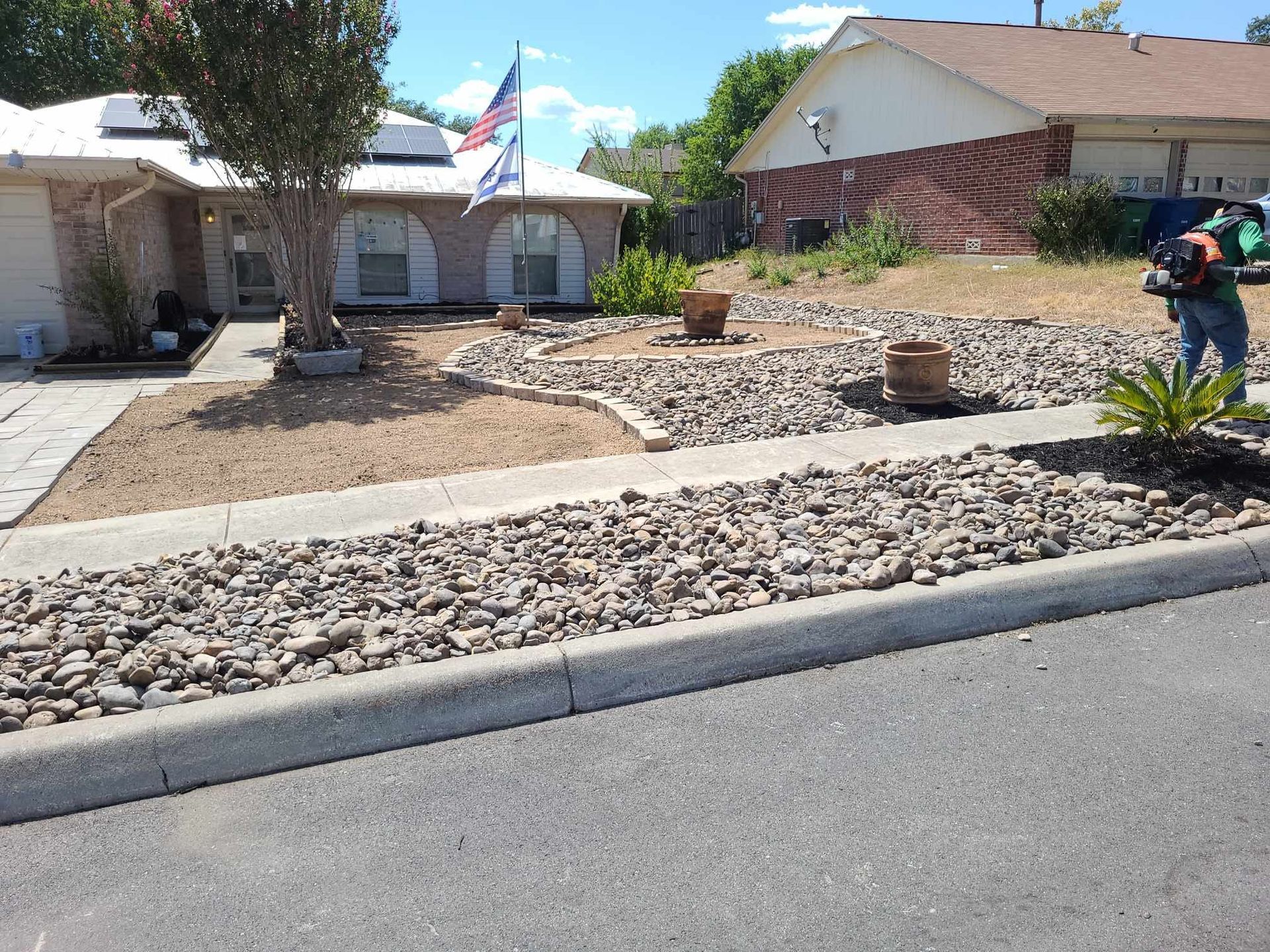 Front yard with rock landscaping and a house, with a person standing on the right holding a hose.