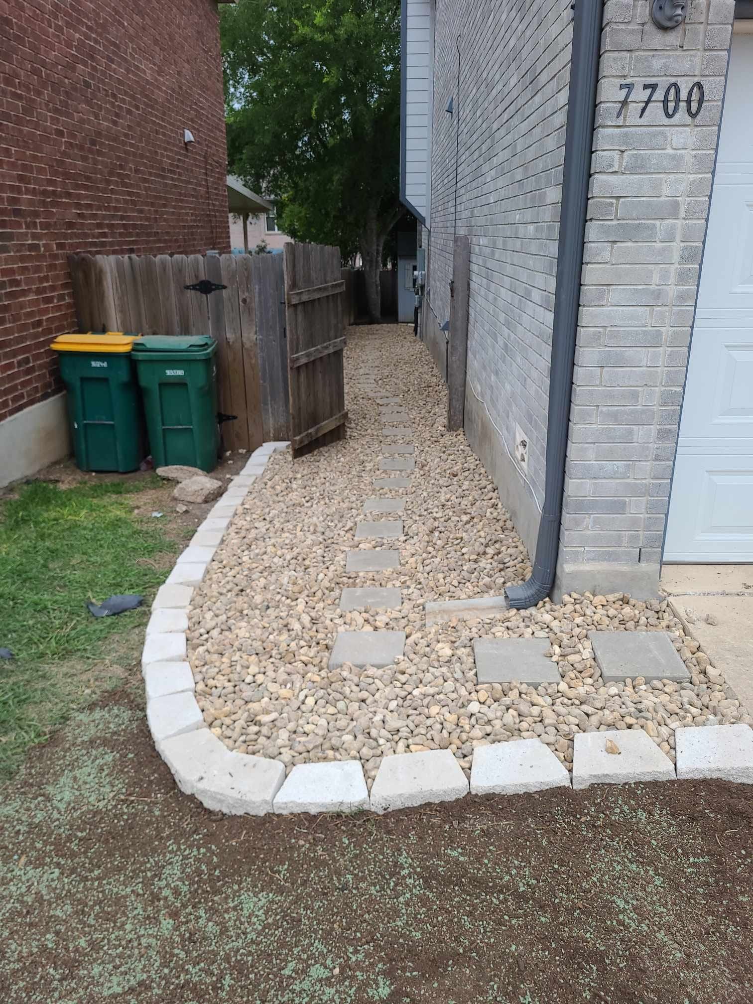 Narrow gravel side path beside brick buildings, with trash bins and a wooden fence near a narrow passage.
