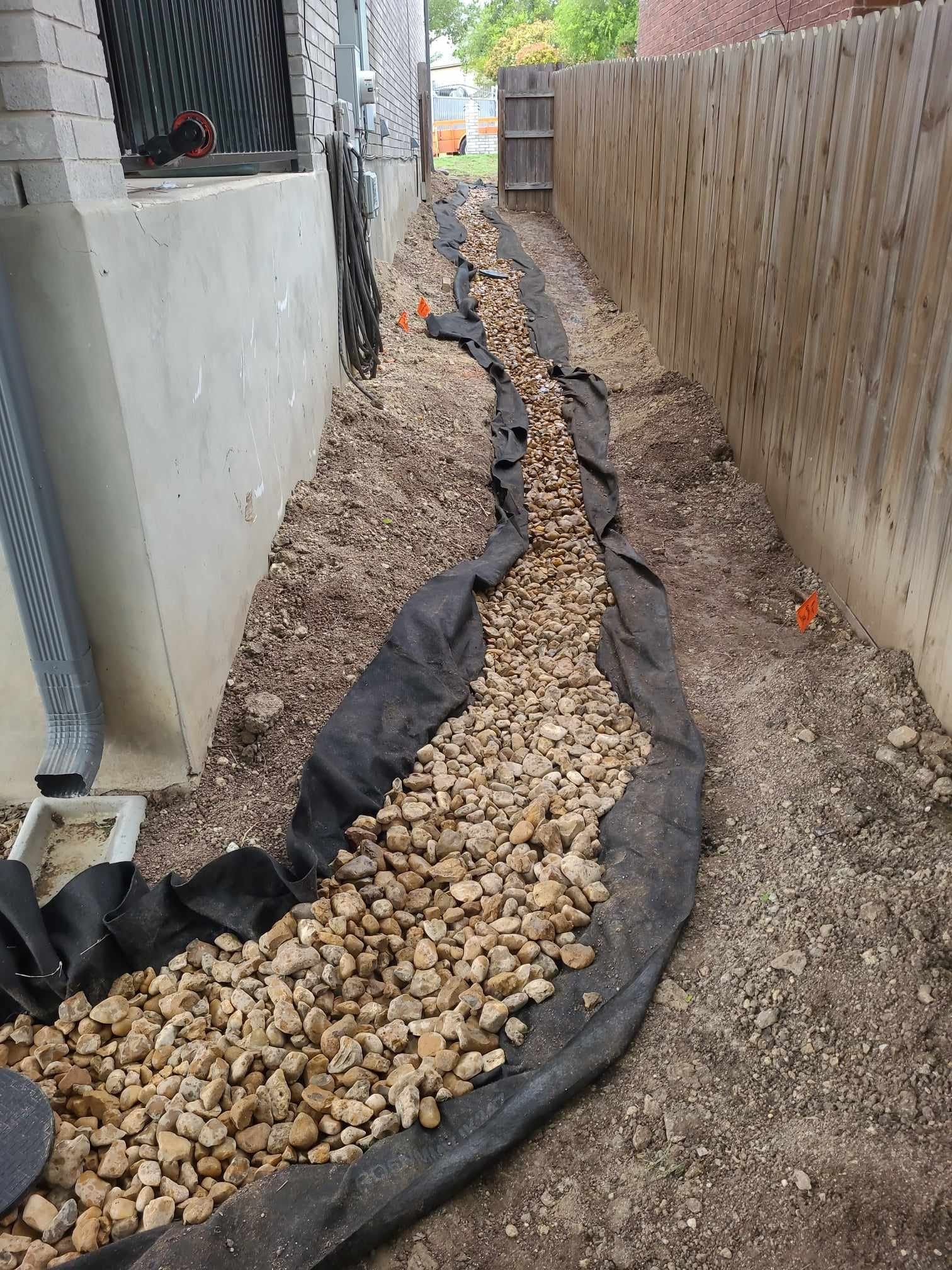 Narrow gravel drainage trench beside a house and wooden fence, lined with black fabric.